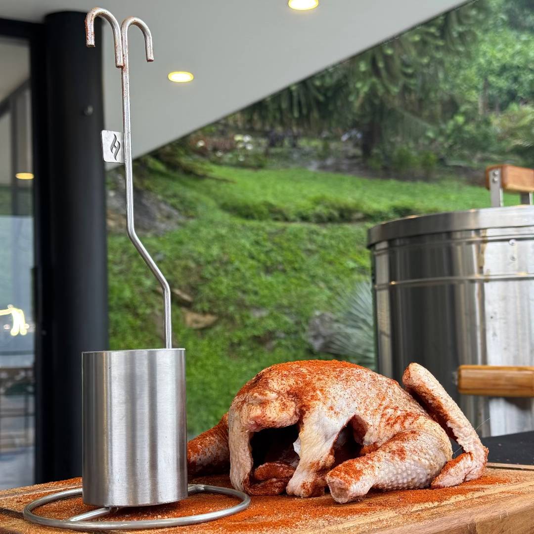 accesorio para pollo sobre tabla de madera y al lado un poco sazonado en paprika con un barril asador de fondo